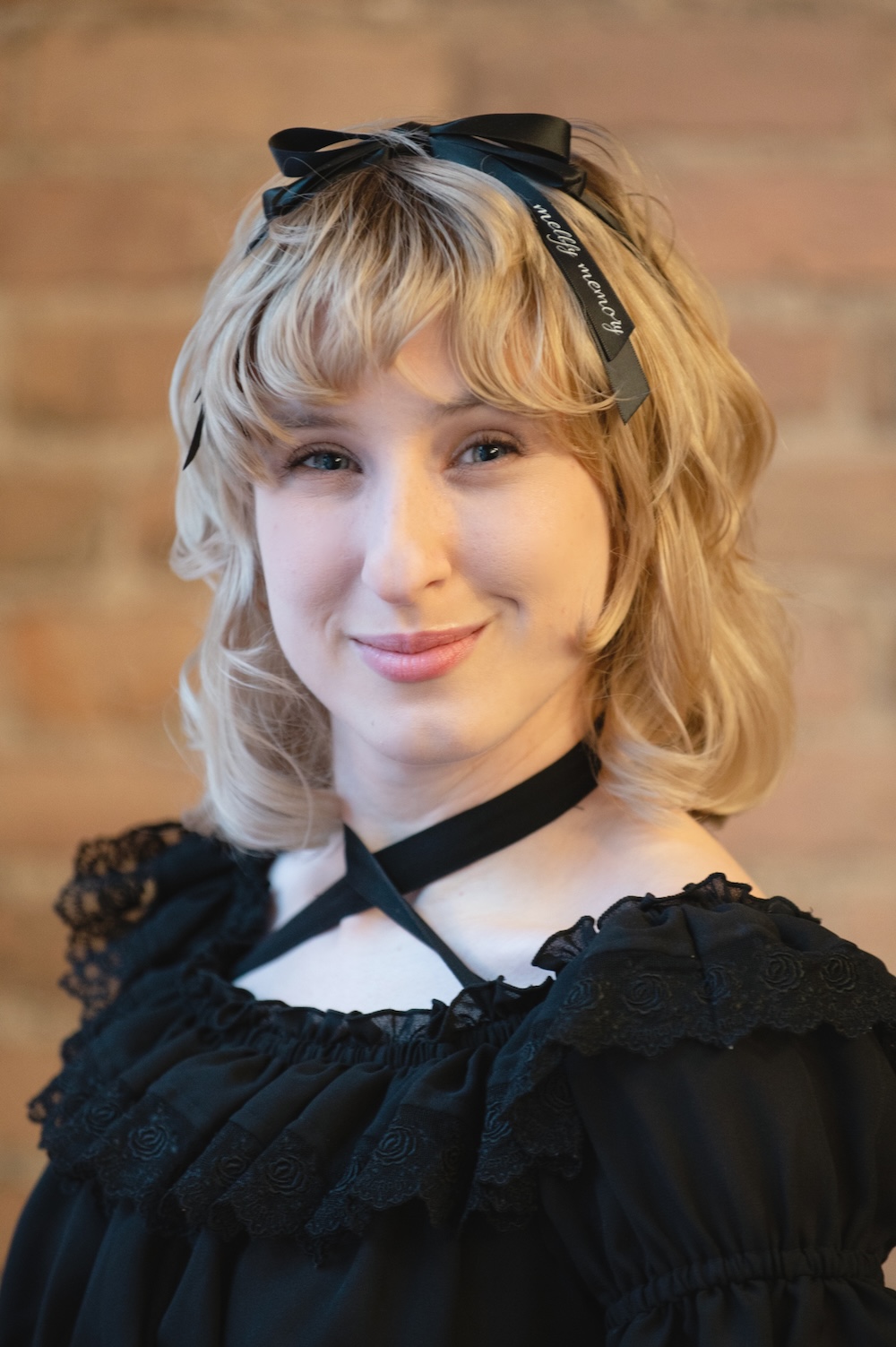 Portrait of a woman with blonde hair wearing a black lace blouse and a ribbon headband, smiling softly.