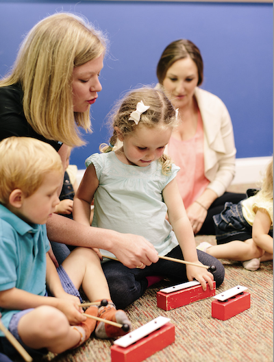 Music Playhouse small - ACM School of Music Teacher guides a young girl tapping a red xylophone with a mallet in a classroom setting. Other children sit nearby, engaging in music activity.